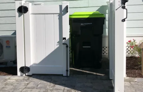 A white, roofless, vinyl PVC enclosure with black locking hardware. One door is swung open to show the garbage can it is designed to protect.